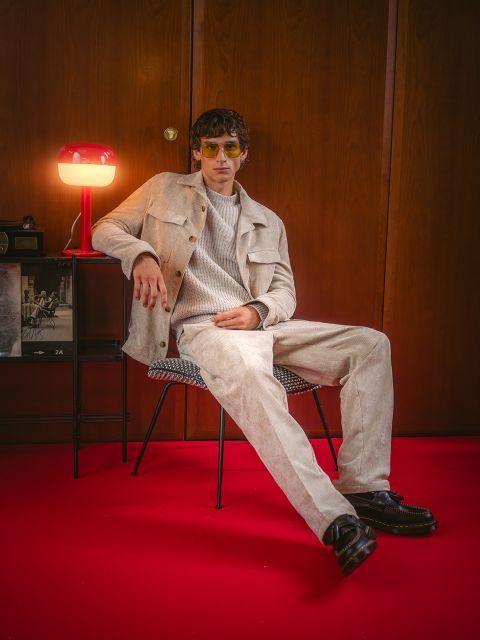 Un hombre con ropa beige está sentado en una silla sobre una alfombra roja, detrás de él hay una lámpara roja y una pared de madera. Lleva gafas de sol y zapatos negros.