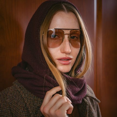 Una mujer con gafas de sol grandes, una capucha de bufanda color burdeos y un abrigo a cuadros marrón posa frente a un fondo de madera.