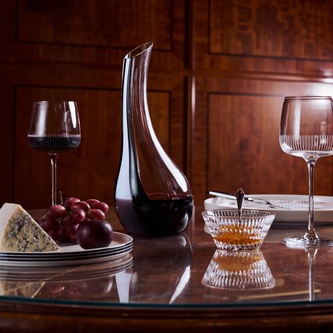 Una jarra de vino, dos copas, queso, uvas y un cuenco de miel sobre una mesa de madera pulida, con platos al fondo.