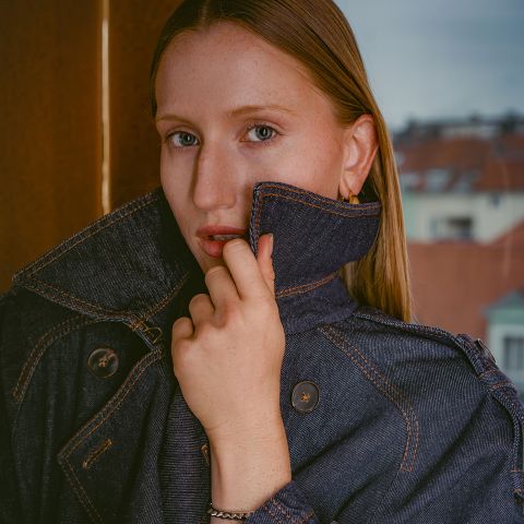 Una mujer de piel clara y cabello liso, que lleva una chaqueta vaquera oscura con el cuello levantado, está de pie frente a una ventana, con edificios visibles al fondo.