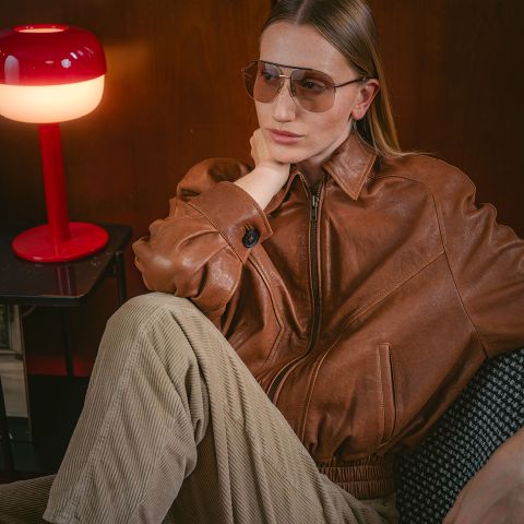 Una persona con una chaqueta de cuero marrón y gafas de aviador está sentada junto a una lámpara roja, con aspecto pensativo, mientras su mano descansa sobre su nuca.
