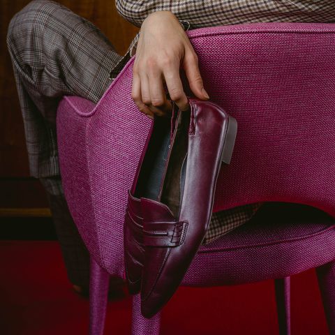 Una persona con pantalones a cuadros está sentada en una silla rosa, sosteniendo un mocasín burdeos sobre el respaldo de la silla.