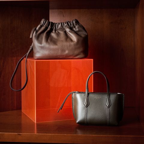 Dos elegantes bolsos de cuero, uno marrón oscuro y otro de color oliva, presentados en un estante de madera junto con un cubo translúcido de color naranja.