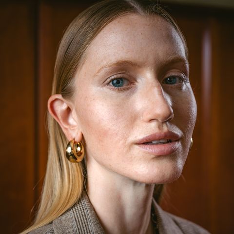 Una mujer con cabello rubio liso, aretes de aro dorados y maquillaje sutil mira a la cámara mientras está de pie frente a un fondo de madera.