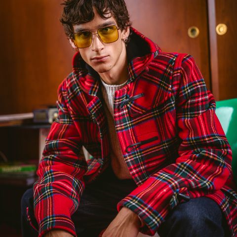 Un hombre con gafas de color amarillo y una chaqueta a cuadros roja está sentado en una habitación, mirando directamente a la cámara con una expresión seria.