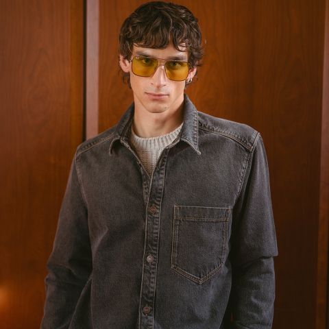 Un joven con cabello castaño ondulado, que lleva gafas tintadas de amarillo, una camisa vaquera gris y un suéter blanco, de pie frente a un fondo con paneles de madera.