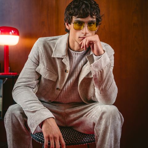 Un hombre con un traje de pana beige y gafas de sol amarillas está sentado en una silla posando pensativamente, mientras que una lámpara roja está sobre una mesa auxiliar junto a él.