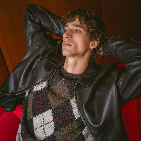 Una persona con chaqueta de cuero y jersey de rombos se reclina en un sillón rojo, con las manos entrelazadas detrás de la cabeza, mirando pensativamente hacia arriba.
