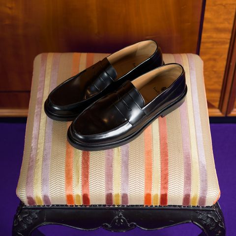 Un par de mocasines negros brillantes descansan sobre un taburete acolchado a rayas en tonos naranja, morado y beige.