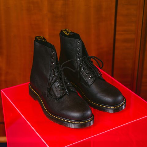 Un par de botas de cuero negras con cordones y costuras decorativas amarillas se exhiben en un brillante soporte rojo frente a un fondo de madera.
