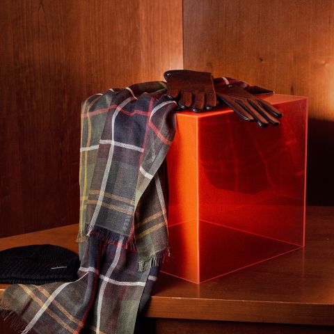 Una bufanda a cuadros y guantes de cuero marrón yacen sobre un cubo rojo translúcido, junto a un gorro de punto negro sobre una base de madera.