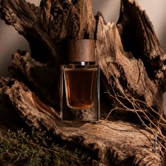 Un frasco de perfume de cristal con un tapón de madera se encuentra en medio de corteza de árbol áspera y ramas secas, transmitiendo así una atmósfera rústica y natural.