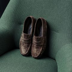 Un par de mocasines de ante marrón, apoyados en un sillón verde texturizado.