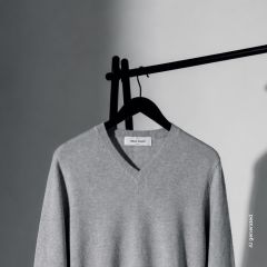Un suéter gris claro con cuello en V cuelga de una percha negra frente a un fondo liso, en parte en sombra, en parte en luz.