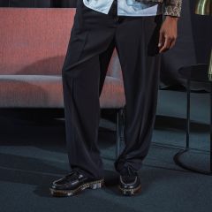 Una persona está de pie en un interior y lleva pantalones negros de pierna ancha y zapatos de plataforma negros; junto a ella hay un sofá rosa y una pequeña mesa redonda.