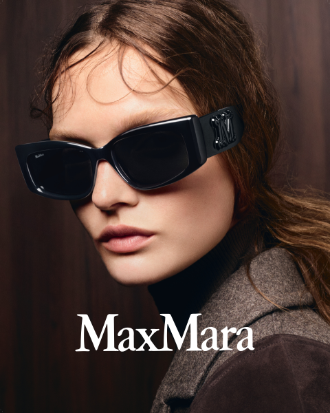 Una mujer con grandes gafas de sol negras y un abrigo marrón delante de un fondo de madera oscura, con el logotipo de "MaxMara" visible en la parte inferior.