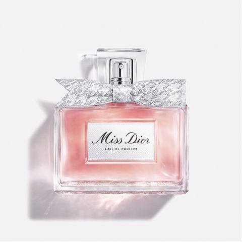 Una botella de cristal transparente de "Miss Dior Eau de Parfum", llena de líquido rosa y con un lazo plateado estampado alrededor del cuello de la botella, sobre un fondo blanco liso.