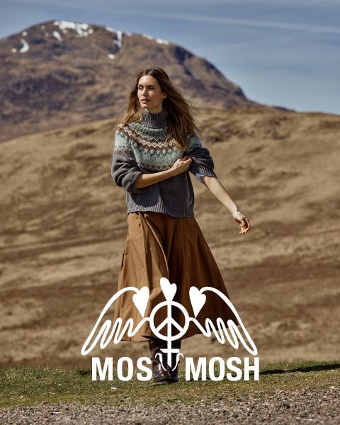 Una mujer con un jersey estampado y una falda marrón está de pie al aire libre, con colinas y una montaña detrás de ella. El logotipo de "MOSS MOSH" es visible en la parte inferior de la imagen.
