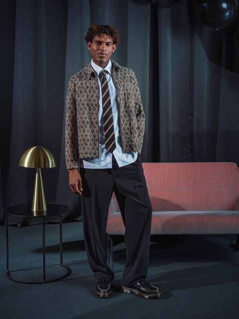 Un hombre con una chaqueta estampada, corbata y pantalones de traje está de pie junto a un sofá rosa y una lámpara dorada frente a cortinas oscuras.