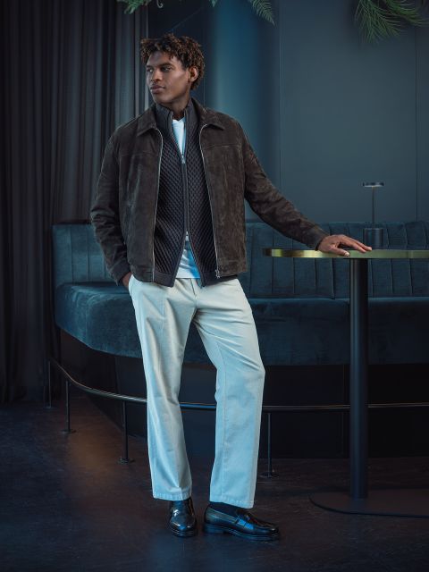 Un hombre con una chaqueta marrón y unos pantalones blancos está de pie junto a una mesa redonda en un salón moderno, tenuemente iluminado, con asientos azul oscuro y cortinas.