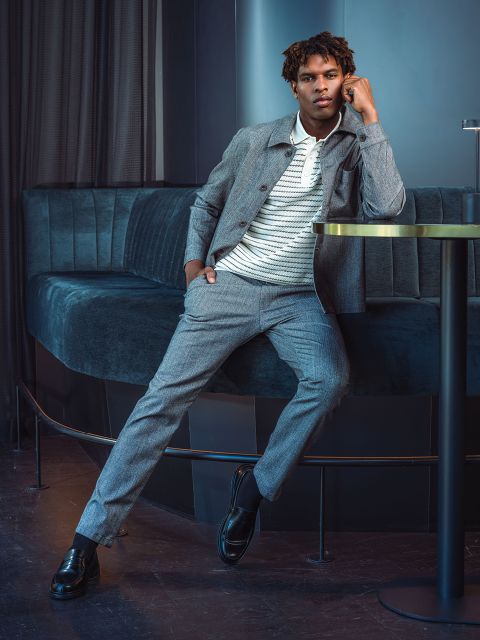 Un hombre con un traje gris y una camisa a rayas se apoya en una mesa redonda en un salón moderno con sillones de terciopelo azul oscuro.
