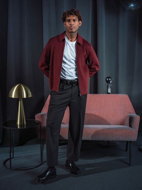 Un hombre con una chaqueta burdeos y pantalones negros está de pie junto a un sofá rosa, sobre el cual hay una mesa redonda con una lámpara dorada, frente a cortinas oscuras.