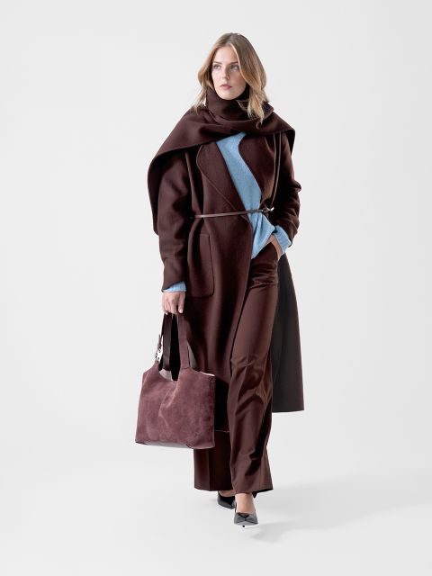 Una mujer con un abrigo marrón oscuro y unos pantalones marrón oscuro, además de un suéter azul, sosteniendo un bolso marrón a juego en la mano y caminando con confianza frente a un fondo blanco liso.