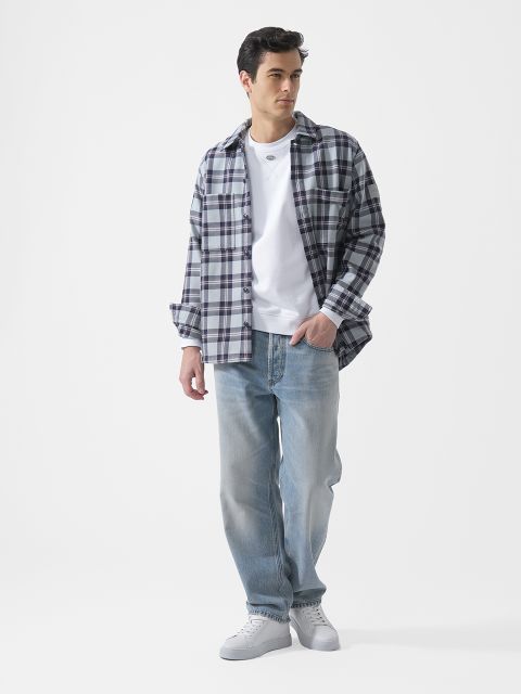 Un hombre que lleva una camiseta blanca, una camisa a cuadros, vaqueros azul claro y zapatillas blancas está de pie frente a un fondo blanco liso.