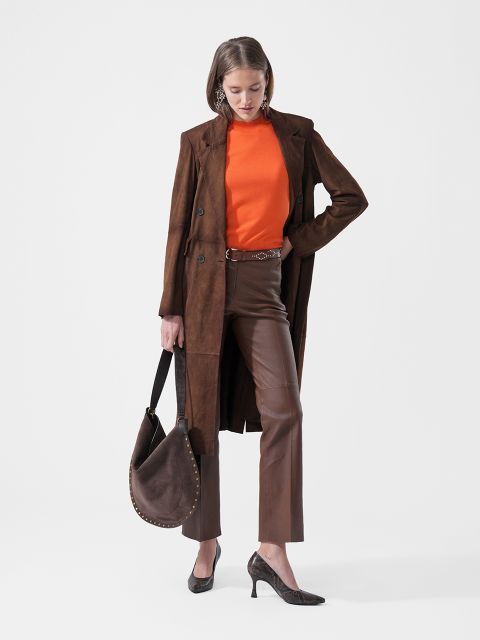 Una mujer con un abrigo marrón, una blusa naranja, pantalones marrones y zapatos de tacón sostiene un bolso de mano marrón y está de pie frente a un fondo blanco liso.