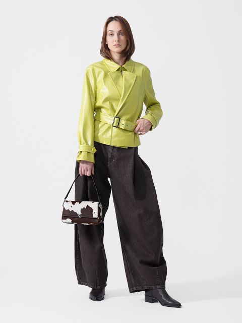 Una mujer está de pie con una chaqueta verde lima con cinturón, unos vaqueros azul oscuro de pierna ancha y botas negras, y sostiene un pequeño bolso de mano con estampado de vaca.
