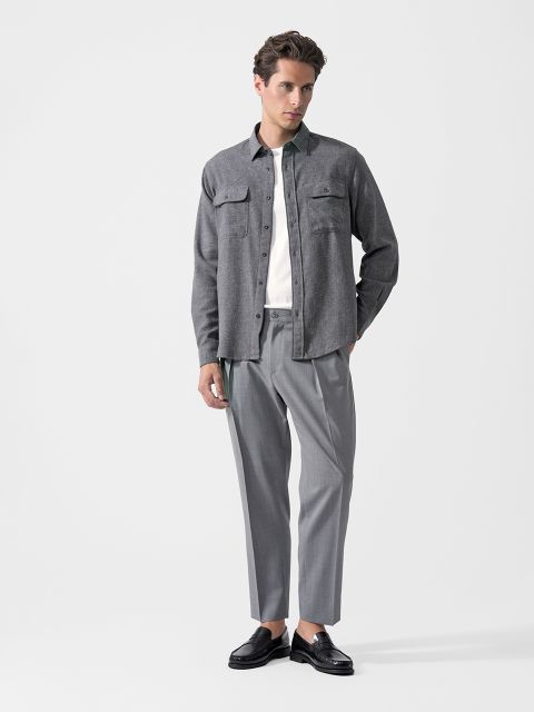Un hombre que lleva una camisa gris con botones, una camiseta blanca, unos pantalones de traje grises y mocasines negros, y está de pie frente a un fondo blanco liso.