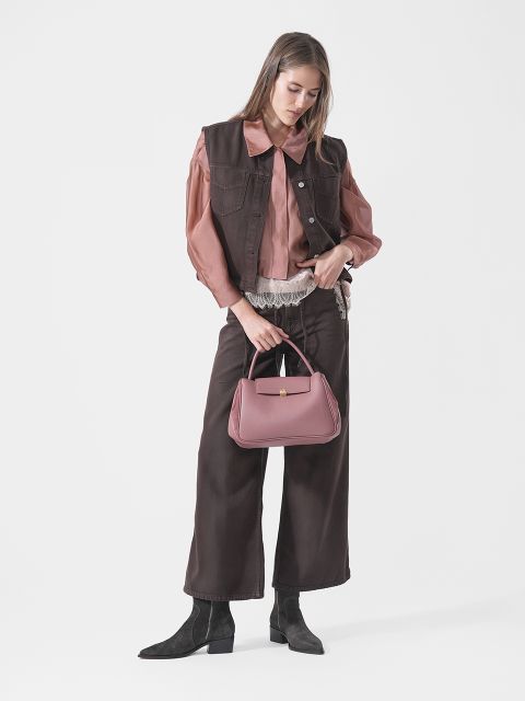Una mujer con chaleco y pantalones marrones, blusa rosa y botas negras sostiene un bolso rosa y está de pie frente a un fondo blanco liso.