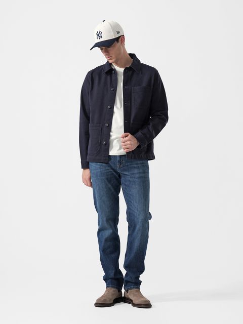 Un hombre que lleva una chaqueta azul marino, una camiseta blanca, vaqueros azules, botas grises y una gorra de béisbol blanca de NY, está de pie frente a un fondo blanco liso.