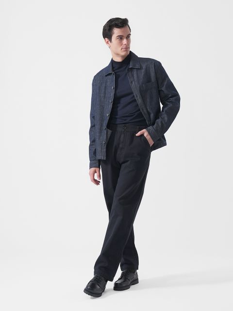Un hombre que lleva una chaqueta vaquera oscura, un jersey de cuello alto azul marino, pantalones negros y zapatos negros, está de pie frente a un fondo blanco liso y mira hacia la izquierda.