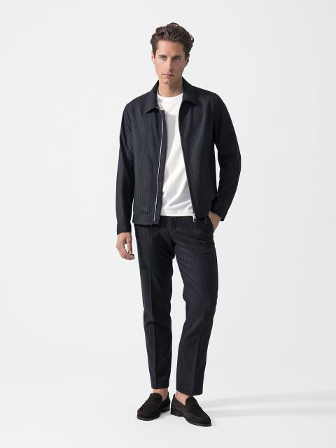 Sobre un fondo blanco, un hombre está de pie, vistiendo una chaqueta oscura de rayas diplomáticas, pantalones a juego, una camiseta blanca y mocasines negros.