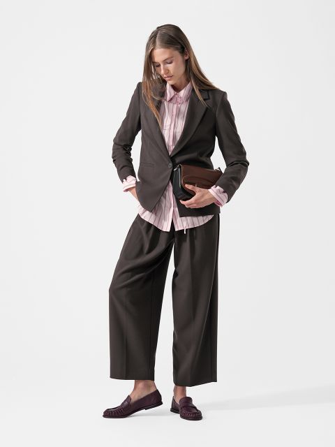 Una mujer con un traje de pantalón oscuro, una camisa rosa y mocasines sostiene un bolso de mano marrón, de pie frente a un fondo blanco liso y mirando hacia abajo.