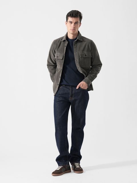 Un hombre con una chaqueta gris, jeans azul oscuro y zapatillas marrones está de pie frente a un fondo blanco liso, con las manos en los bolsillos.