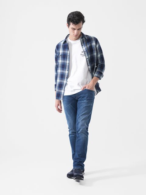 Un hombre con una camisa de cuadros azul, una camiseta blanca, vaqueros azules y zapatillas, caminando frente a un fondo blanco liso y mirando hacia abajo.