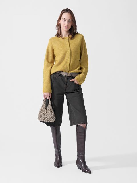 Una mujer que lleva un suéter amarillo, pantalones cortos negros hasta la rodilla y botas marrones, y sostiene un bolso trenzado en la mano, está de pie frente a un fondo blanco.
