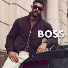 Un hombre con una chaqueta marrón, gafas de sol y pantalones claros se apoya en un coche junto a la inscripción "BOSS".