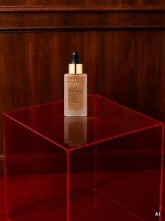 Sobre una mesa de acrílico rojo transparente, frente a una pared de madera y una alfombra roja, hay una botella de suero Estée Lauder.