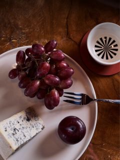 Un plato con uvas rojas, una rebanada de queso azul y una ciruela; también se ven un tenedor y una taza sobre una mesa de madera.