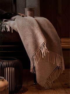 Una manta beige con flecos colocada sobre una silla, junto a una vela en un farol negro y una taza de café en el suelo de madera.