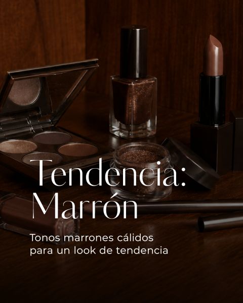 Productos de maquillaje marrones, incluyendo sombra de ojos, lápiz labial, esmalte de uñas y brochas, sobre una superficie de madera con texto en español sobre la tendencia del marrón.