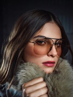 Una mujer de pelo largo y castaño lleva gafas de aviador tintadas y un abrigo con cuello de piel, mirando a la cámara con confianza sobre un fondo oscuro.