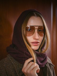 Una mujer con cabello largo, que lleva gafas de aviador, tiene una bufanda marrón sobre la cabeza y lleva un abrigo de cuadros marrón, está de pie frente a una pared de madera.