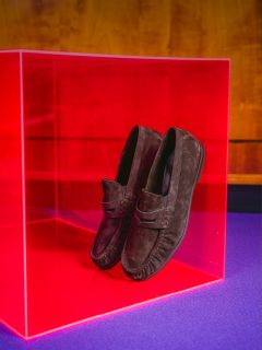 Mocasines de ante marrón, presentados en un cubo rosa transparente, sobre un fondo de madera y sobre una base morada.