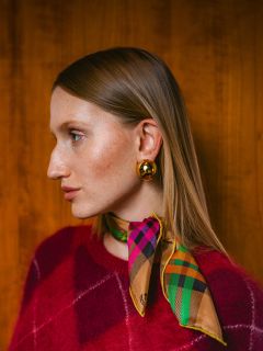 Mujer con un suéter rojo, una bufanda a cuadros de colores y aretes dorados, mirando hacia la izquierda, frente a un fondo de madera.
