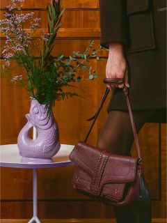 Una persona con ropa oscura sostiene un bolso burdeos junto a un jarrón morado con forma de pez con flores sobre una mesa blanca.