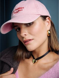 Una mujer con cabello castaño liso, gorro rosa, aretes dorados, gargantilla de corazón y top rosa peludo, mira a la cámara frente a un fondo azul.